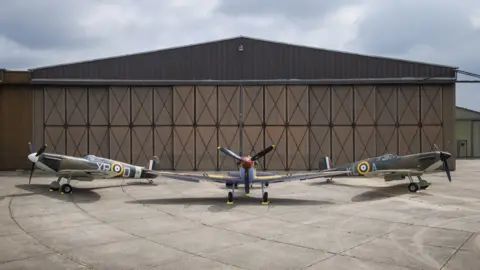 IWM Three Spitfires (l-r Mk Ia X4650, Mk V JG891, Mk Ia R213) in front of a hangar at IWM Duxford