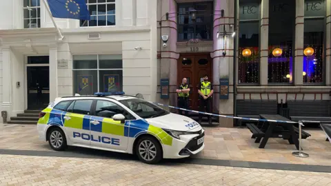 A police car is parked diagonally across a kerb outside a bar in a city street. There are black wooden benches outside obstructed by police tape, and two officers standing outside the ornate wooden doors of the building.
