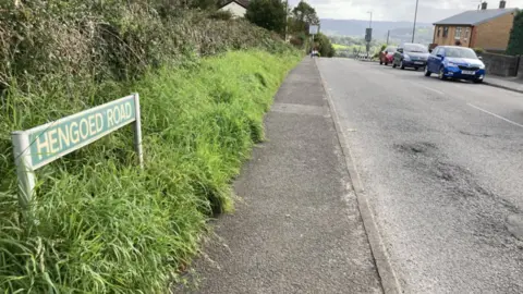 BBC A shot of Hengoed Road, where the incident happened. The road sign is visible as well as a street lined with grass hedges on the left side, and cars and houses on the right side.