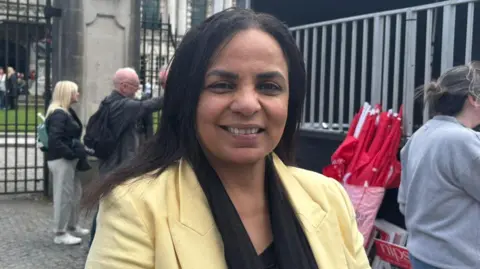 Manal is smiling astatine the camera. She has agelong achromatic hairsbreadth and brownish skin. She is wearing a achromatic blouse and yellowish blazer. She is stading successful beforehand of the gates of Belfast City Hall.