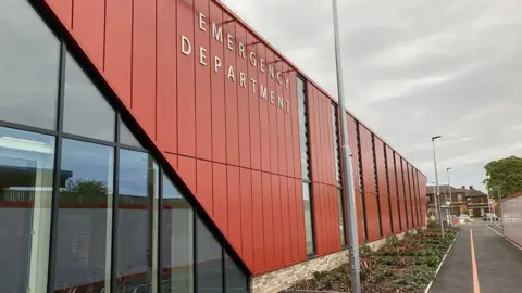 Red and glass building with Emergency Department written on it. There are plants, a pavement, lamp posts and a fence.

There are houses in the background.