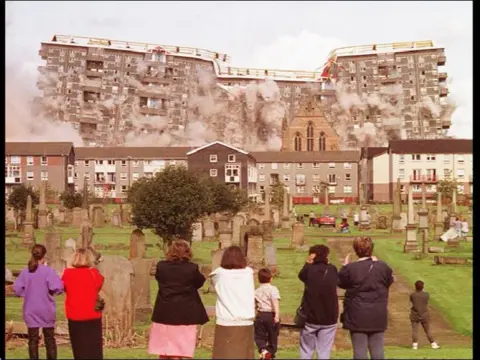 PA Media A head-on image of a row of high rise flats, mid-explosion. We look on from behind a row of people watching the whole thing. The flats are collapsing in in themselves from the middle out, plumes of dust going into the sky.