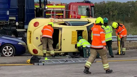 BBC A yellow car on its side in front of a fire truck with five men attending it
