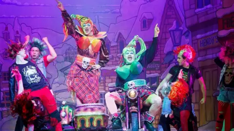 The Grove Theatre Six actors on stage, dressed in colourful costumes as punks. One is sat on a motorbike, with one standing on a seat next to him. The two in the middle have their arms out. Stage scenery is behind them. 