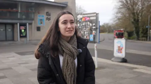 Young womanwith dark hair wearing coat and long scarf standing in front of a concrete building