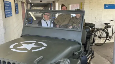 A 1940s green jeep with historical reenactors inside a straight brick corridor with a militaristic feel.