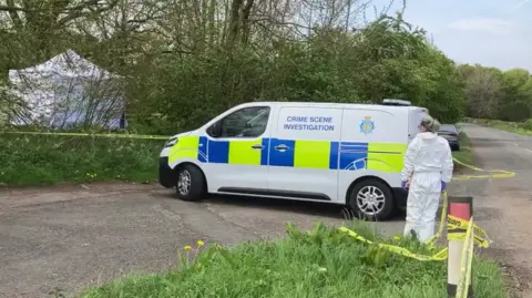 A police van with branding on it that reads: "Crime Scene Investigation" is parked outside an area of woodland. A white tent can be seen in the distance and a police cordon surrounds it. Someone in a white hazmat suit is walking towards the van.