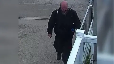 Lincolnshire Police A man wearing a dark green bomber jacket, black cargo trousers and a black backpack. He is bald with a beard and is walking with his head facing the ground. There is a white fence to the right of the image. 