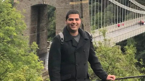 Itamar Bareket, a man with dark hair and a bright smile, he wears a black formal coat and a rucksack, with stands in front of a grand bridge.
