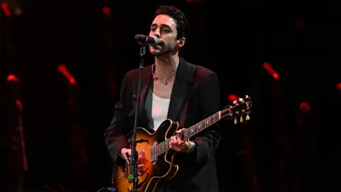 Getty Images Stephen Sanchez performs at Glastonbury. He's wearing a black blazer with a white vest underneath and a necklace, singing into the microphone. He is holding a guitar which is coloured orange and black. The background has red coloured staging.