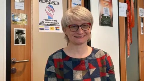 A woman with silver hair in a bob and a multi-coloured top sits in a chair, smiling at the camera. The wall behind her is decorated with pictures and posters, including one which reads: "TRANS RIGHTS ARE HUMAN RIGHTS".