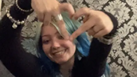 A young girl with long blue hair holds up a heart sign with her hands. She is smiling and wearing a long sleeved black top, silver bracelets. She is sat in front of black and silver patterned wallpaper.