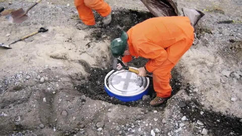 USGS/Science Photo Library A scientist in an orange jumpsuit placing a round seismometer in a prepared hole in the ground