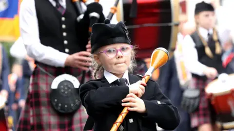 Pacemaker ARCHIVE PHOTO: A young girl parading in Gilford, County Down, last year. She is wearing pink-rimmed glasses, a black ceremonial jacket and hat, a white shirt and a patterned tie. She is holding a wooden baton which can be twirled during a march.