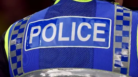 A close-up of the back of a police officer who is wearing a blue hi-vis vest with the word 'police' written in white letters. There are two reflective strips down either side in a grey and blue grid pattern.
