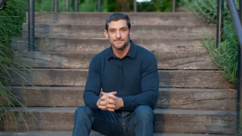 Shira H. Weiss Amir sits on wide wooden outdoor steps, leaning slightly forward with his hands clasped together. He is wearing a dark long‑sleeve top and jeans.