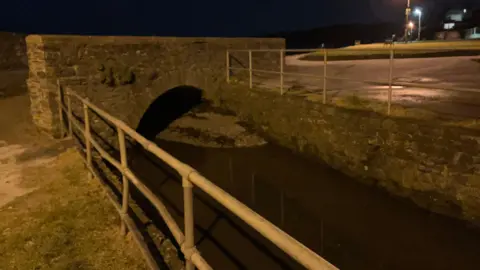 A river with metal railings on either side and a stone bridge over it.