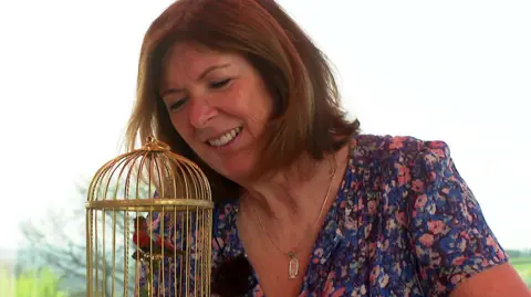 A woman with short brown hair wearing a necklace and blue floral top. In front of her is a small red bird with blue and green accents on its feathers inside a golden cage. The background is white and green. 