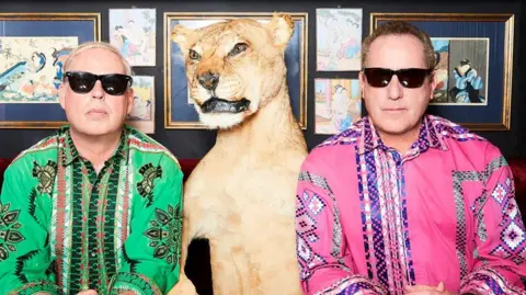 Two men wearing black sunglasses are pulling serious poses whilst wearing very colourful shirts with patterns/ One man has short blond hair and a green shirt and the other on the right has a pink shirt and short brown hair. In the middle is a taxidermy lion and there are photos on the wall behind them.
