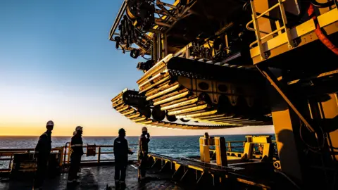 The Metals Company The crew of a ship look up at the tracks of a subsea mining vehicle.