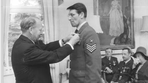 Bolton Family A black and white photo of a man in a suit pinning a medal on the chest of Geoffrey Bolton in 1972. Flt Lt Bolton has three stripes on his shoulder and there are further RAF personal sitting and a woman wearing a hat behind him in a room with a painting on the wall.