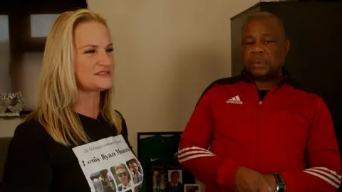 UKFS Cheri Curran, standing next to Rohan Shand Snr, in a film. They are in a room. She has a black T-shirt on with her son's name on it and images of him. Rohan is wearing a red Adidas tracksuit top and is looking down. Cheri is looking to the right. There is a mirror and photos behind them. 