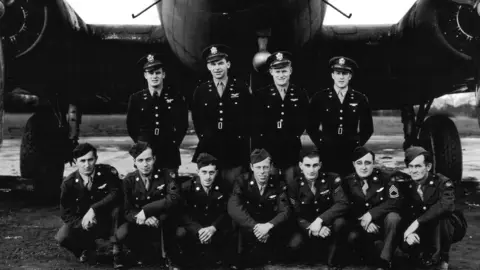 384th Bombardment Group Crew members in uniform in front of their plane. There are seven kneeling down in the front row and four more standing behind. It is a black and white image. 