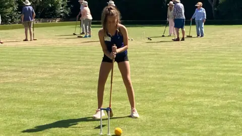 A teenage girl dressed in a blue vest and black shorts is swinging her mallet to hit a ball through a hoop. In the distance are other croquet players.
