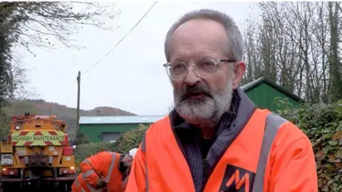 Shropshire Council A man wearing an orange hi-vis jacket and glasses pictured from the shoulders up, with another man in hi-vis behind him and a highway maintenance vehicle behind.