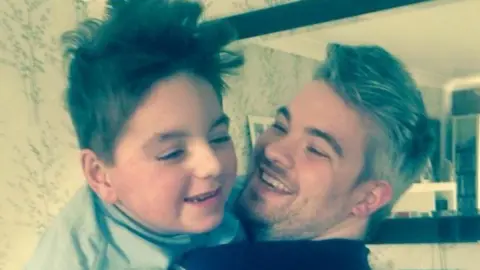 Paul Woodward Paul Woodward his holding his son Kalel in his arms and they are both laughing. They are standing in front of a mirror in a living room.