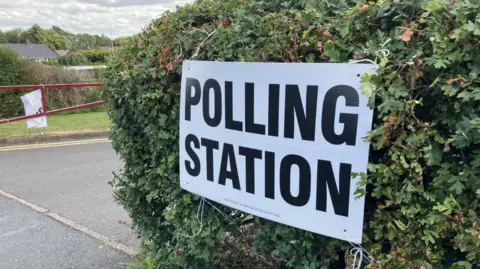 A white sign attached to a hedge reads POLLING STATION in blocked capital black lettering.