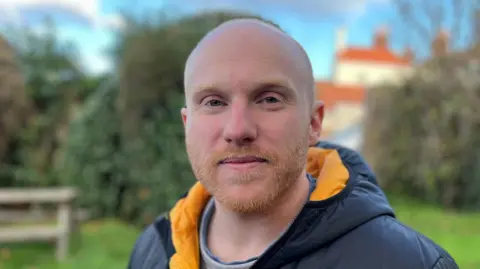 BBC A man looking slightly side on at the camera smiling wearing a black and yellow puffer coat standing in a field.