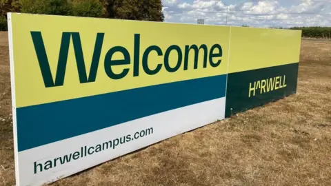 A sign reads "Welcome, Harwell". It is yellow, blue, white and black and positioned on the ground.