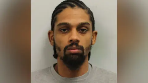 A man in a grey shirt with a beard. He looks at the camera in a mugshot image.