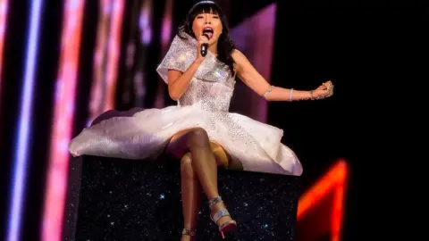 Getty Images Singer Dami Im sits on a glittery box in a sparkly white dress with her legs crossed