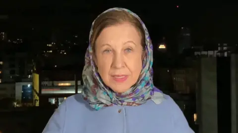 Lyse Doucet in a blue coat and headscarf, she is stood with a dark Tehran skyline behind her