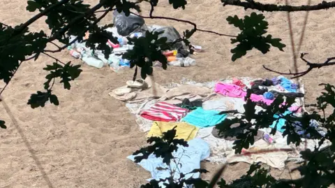 BBC/Phil McCann Several towels remain on a sandy section by the river, including a pink and white stripey towel, a yellow one, blue one, pink one. Rubbish bags and empty bottles next to them.