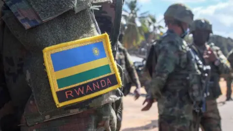 A close-up of a soldier’s uniform showing a Rwandan flag patch on the shoulder. Armed soldiers stand in the background.
