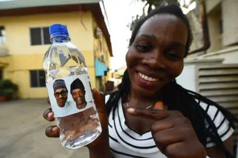 AFP A woman smiles as she holds a bottle of water branded with the faces of President Muhammadu Buhari and the vice-president in Abuja, Nigeria - Wednesday 20 February 2019