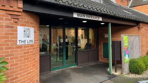 Jo Thewlis/BBC The entrance to The Link at Breckland Business Centre. The entrance door is made of sliding glass and is surrounded by red-brick walls. A sign saying 'The Link' is attached to the wall to the left hand side of the doors.