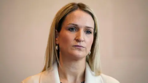 PA Media Helen McEntee - A woman with blonde, shoulder-length hair wears earrings and a white suit jacket. She is standing in front of a blank, neutral colour wall.