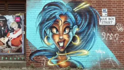Trik09/Instagram A spray painting on a brick wall. A woman with big cartoon eyes smiled at the camera. She has red lipstick on and a gold necklace which is semi-floating, and matching hoop earrings, a hair accessory and cuff ear piercing.