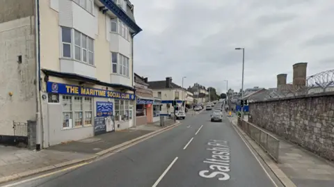 Google Google street view of The Maritime Social Club on Saltash Road in Plymouth. The four-storey building has two bay windows on the mid two floors, below there is a blue sign with yellow writing saying 'The Maritime Social Club' and other signs with writing that cannot be made out. Parked and driving cars are visible in the background. Overcast sky.