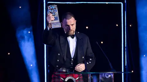 Getty Images A man in a kilt receives an award trophy