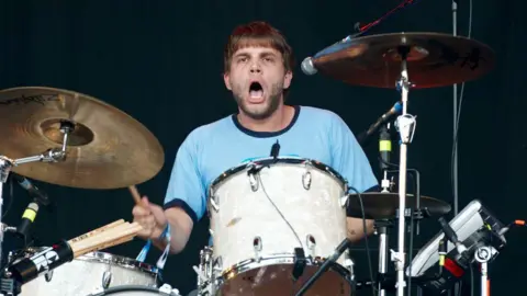 Getty Images A man in a blue T-shirt with a beard and brown hair plays a white drum with cymbals beside it