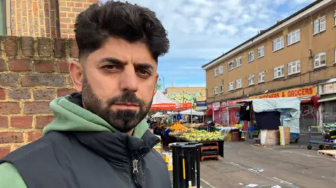 BBC Adam Kadirzadah in a green hoodie and black gilet. He has dark hair and a beard and moustache. 