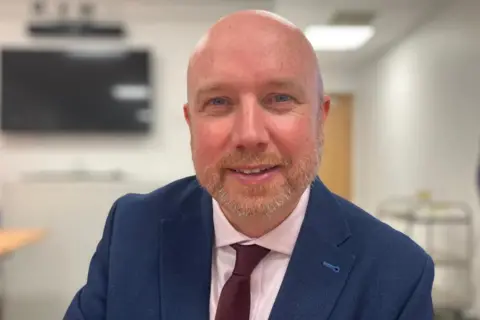 A head shot of a bald man smiling. He is wearing a blue suit jacket with a white shirt and a maroon tie.