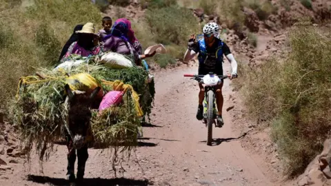 AFP A competitor waves at a Moroccan woman as he rides his bikes during the 600 km ( 372 miles) Titan Desert mountain bike race on 30 April 2018