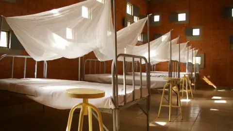 Francis Kéré Centre for Health and Social Welfare. There are beds lines up with white nets covering them. The walls are brick and there is light entering from small windows.