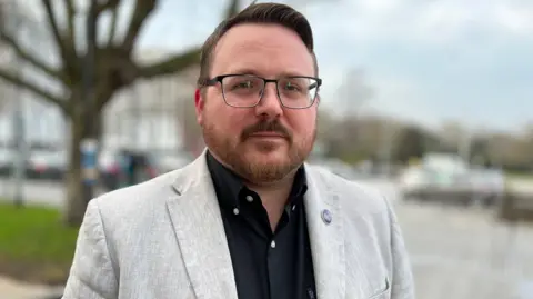 A man in a cream-coloured blazer and black shirt with glasses and beard looks at the camera.